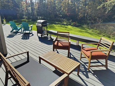A backyard deck with wooden furniture, two blue lounge chairs, a barbecue grill, and a sunny lawn in the background.