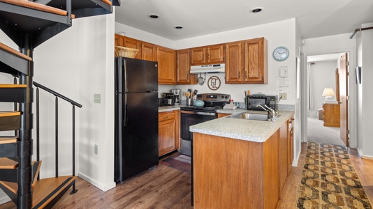 A compact kitchen with wooden cabinets, a black refrigerator, stove, microwave, and a small island with a sink. Adjacent spiral staircase and hallway leading to a room with a bedside lamp visible.