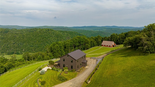 Aerial view of a brown farmhouse and a red barn situated in a lush, hilly landscape with a curving gravel road and surrounding greenery.