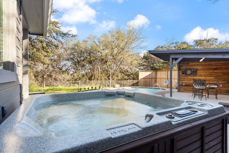 Outdoor patio area with a hot tub in the foreground and a covered seating area with a table and chairs in the background. Trees and a fence are visible in the distance.