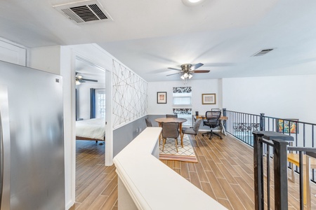 Bright open area with wood flooring, featuring a bedroom, dining space with table and chairs, office desk, ceiling fans, and metal railing. White walls and natural light create a spacious atmosphere.