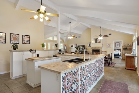 A spacious kitchen with a long tiled island featuring a patterned front, pendant lights, white cabinets, and an adjacent dining area leading to a bedroom. Artwork and decor adorn the beige walls.