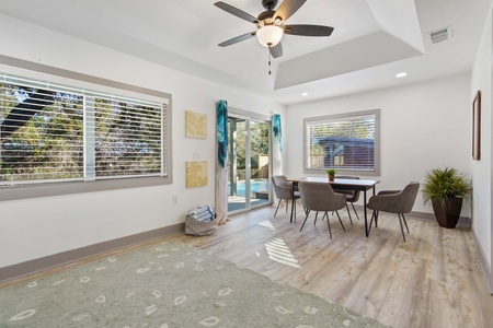 A bright dining area with a table and four chairs, large windows, sliding glass door, ceiling fan, and light wood flooring. There is a green rug and a potted plant in the corner. .