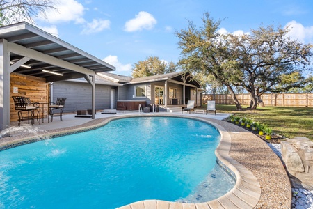 A modern backyard features a swimming pool with a curved design, adjacent to a covered patio area with seating. The yard is landscaped with trees and a fence runs along the perimeter.