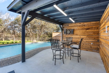 Covered outdoor seating area with a table and four chairs beside a pool. A dartboard is mounted on the wood-paneled wall. Trees are visible in the background beyond a fence.