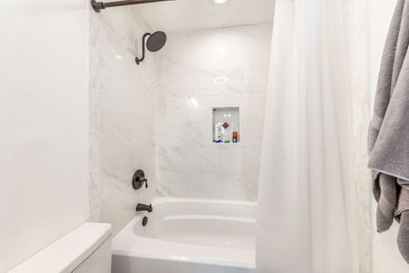 A modern bathroom featuring a white bathtub with a mounted black showerhead and faucet, white marble tiles, a built-in shelf with toiletries, and a light gray towel hanging on the right side.