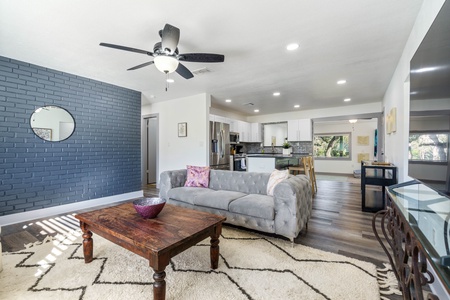 Modern living room with a gray sofa, wooden coffee table, geometric rug, and a dark blue accent wall with a round mirror. Open kitchen in the background with white cabinets and stainless steel appliances.