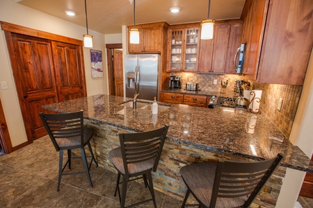 A modern kitchen with granite countertops and 3 high back stools, stainless steel appliances, wooden cabinets, and pendant lighting.