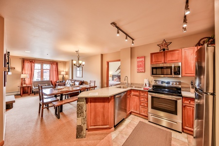 Open-concept kitchen and living area with wooden cabinets, stainless steel appliances, and a granite countertop. The living area features large windows, a wooden dining table, and a cozy seating area.