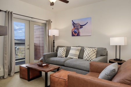 A living room with a sofa, armchair, coffee table, and lamp. French doors open to a balcony with a scenic view.