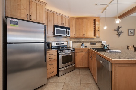 A modern kitchen with stainless steel appliances, wooden cabinets, granite countertops, and tiled flooring. The room is well-lit by pendant lights and features a sink and stove.