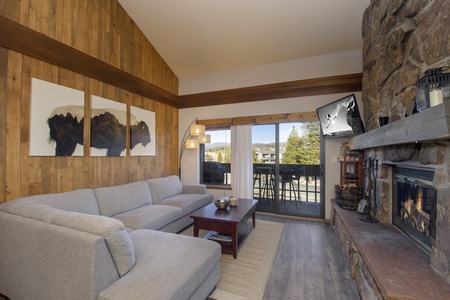 A cozy living room with a gray sectional sofa, wood paneled walls, a fireplace, a large TV,  and a view of trees and mountains through sliding glass doors leading to a balcony.