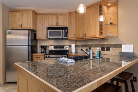 Modern kitchen with wooden cabinets, granite countertops, stainless steel appliances, and an island featuring a double sink and bar stools. An open dish rack, coffee maker, and some cleaning supplies are visible.