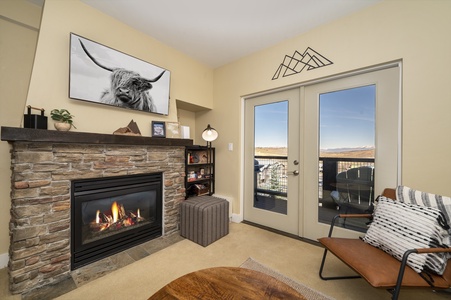 A cozy living room features a stone fireplace, leather chair and double glass doors, bathing the room with natural light, leading to a balcony and an exterior view.