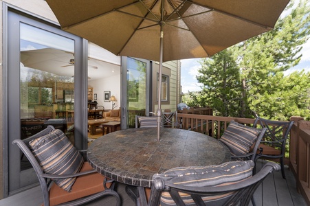 A round stone-topped table with six cushioned chairs and an umbrella is set on a wooden deck, overlooking a lush, green landscape. Large sliding glass doors open to an indoor living and dining area.