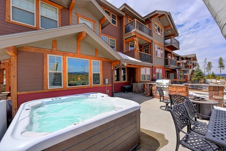 Outdoor private hot tub and and spacious patio with seating area and a gas grill in front of a multi-story wood and stone building on a clear day.