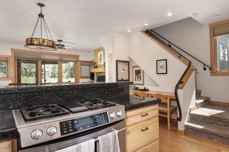 Bright kitchen with a gas stove, granite countertops, and wooden cabinetry. An adjacent staircase leads to an upper floor. Large windows offer natural light and a view of the outdoors.