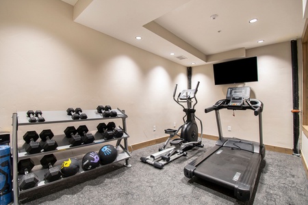Compact fitness room with treadmill, elliptical, free weights, and mounted TV for convenient workouts.