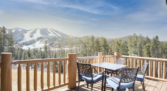 A wooden deck with a table and four chairs overlooks a scenic mountain range with ski slopes and a forest