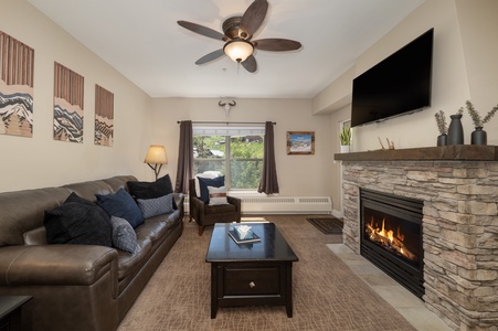 A cozy living room with a leather sofa, coffee table, and stone fireplace. A flat-screen TV is mounted above the fireplace, and artwork decorates the walls. A ceiling fan and lamp provide lighting.