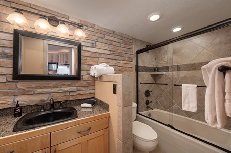 A modern bathroom with a black sink, wooden cabinet, large mirror, and towel rack. The shower has a glass door and is equipped with a showerhead and a towel hanging over the rail. The walls are tiled.