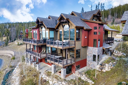 A three-story modern wooden house with multiple balconies is situated on a hillside surrounded by scattered trees