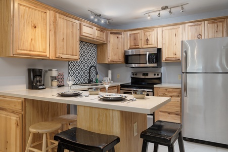 A modern kitchen features light wood cabinets, a stainless steel refrigerator and stove, and a kitchen island with stools.