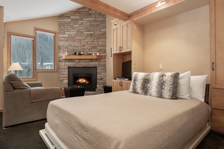 A cozy bedroom with a stone fireplace, a bed with beige bedding and fur pillows, and a brown armchair. The room features a wall-mounted TV, wood accents, and two windows with a snowy view outside.