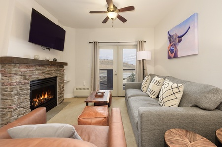 A cozy living room with a modern decor, featuring a sofa, leather chair, fireplace, wall-mounted TV. A glass door and window provide natural light an outdoor view.