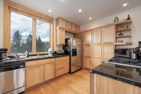 A modern kitchen with light wood cabinets, black countertops, stainless steel appliances, a large window, and recessed lighting.