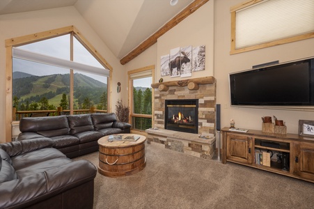 A cozy living room with a leather sofas, rustic wood accents, a stone fireplace, a flat-screen TV, and mountain views through large windows.