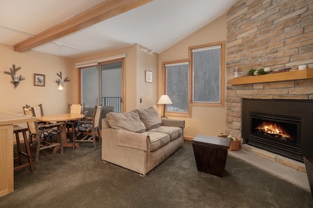 A cozy living room with a stone fireplace, couch, wooden coffee table, and a dining area with a table and chairs. Beige walls and carpet, with large windows and exposed wooden beam on the ceiling.