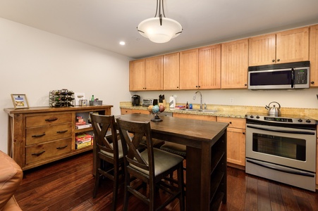 A modern kitchen with wooden cabinets, a stainless steel appliances. A wooden island with four chairs sits in the center. A sideboard with a built-in wine rack.