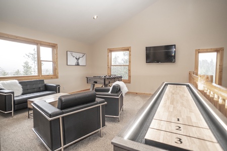 A modern loft-style game room with black leather furniture, a TV on the wall, and a shuffleboard table. The room also has a foosball table and  windows provide natural light.