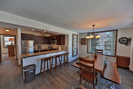 A modern kitchen and dining area with wooden furniture. The kitchen features stainless steel appliances and a bar with three stools. The dining area has a rustic wooden table under a chandelier.