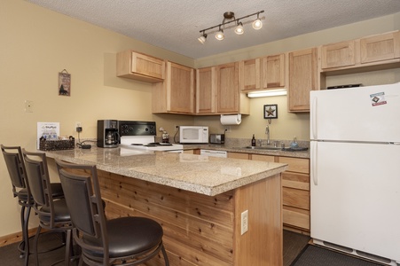 A cozy kitchen with granite counter tops, wooden cabinets, a refrigerator, a stove, a microwave, and a drip coffee maker. .