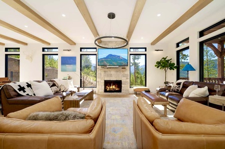 A spacious living room with leather sofas, a fireplace, and a wall-mounted TV. Large windows and wooden ceiling beams enhance the bright, modern decor. A forested view is visible outside.