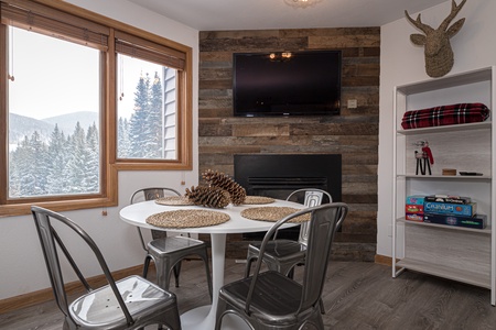 A small dining area with a round table and four metal chairs. The views out the window of the forest and a ski run