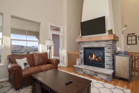 A cozy living room with a leather sofa, stone fireplace, and a mounted TV. A large window offers a view of mountains