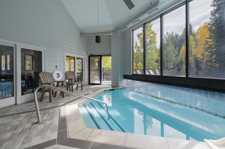 Indoor swimming pool with large windows overlooking a forested area. The space includes cushioned wooden chairs, pool handrails, and natural light filtering through the windows.