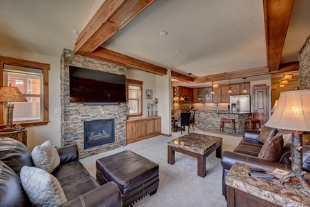 A stylish living room with a modern rustic design featuring leather sofas, stone fireplace, flat-screen TV, wooden beams ceilings, and an open kitchen with bar stools and stainless steel appliances