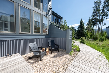 - Enjoy a peaceful moment on this private gravel patio with two modern chairs - Wood stump tables add rustic charm against the sleek gray siding and walls - Surrounded by nature with a wooden path leading to scenic views nearby