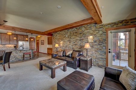A cozy living room with stone accent wall, leather couches, and a coffee table. An open kitchen with bar stools and a glass door leads to an outdoor area.