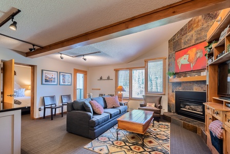 A cozy living room featuring a stone fireplace, sofa, armchair, wooden coffee table, and a colorful area rug. The room has large windows, wooden accents, and an open doorway leading to a bedroom.