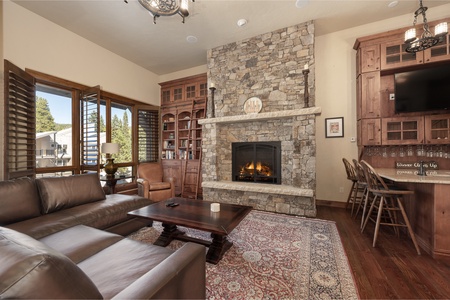 A cozy living room with a stone fireplace, wooden furniture, a leather sectional sofa, and wooden cabinetry. A television is mounted above the fireplace, and a large rug lies in front of the sofa.