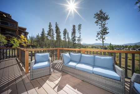 A sunlit outdoor deck with wicker furniture, including a love seat and armchair. The deck overlooks a scenic landscape