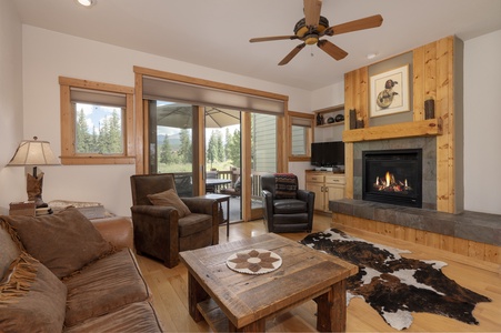 A cozy living room with a gas fireplace, a leather sofa, armchairs, and a wooden coffee table. Sliding glass doors open to a patio with outdoor seating, overlooking a forested landscape.