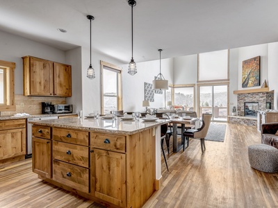 A modern kitchen with wooden cabinets, a large island with granite countertops, pendant lights, open to a dining area, and a living room with a fireplace and large windows bathing the room in natural light