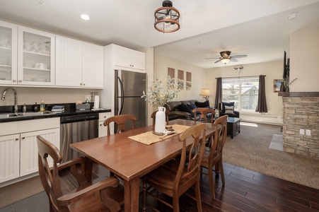 Open-concept kitchen and living area with white cabinets, stainless steel appliances, a wooden dining table, and a cozy seating area with a stone fireplace and ceiling fan.