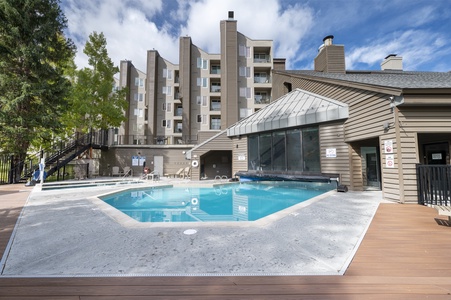 An outdoor pool surrounded by a concrete patio area next to a multi-story residential building with balconies, trees, and a clear blue sky in the background.